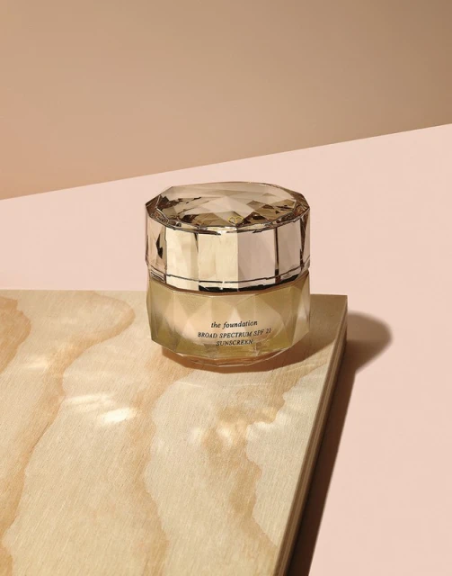 Close-up still life of skincare foundation in a jewel-faceted jar, placed on a light wood surface, against a tan backdrop.
