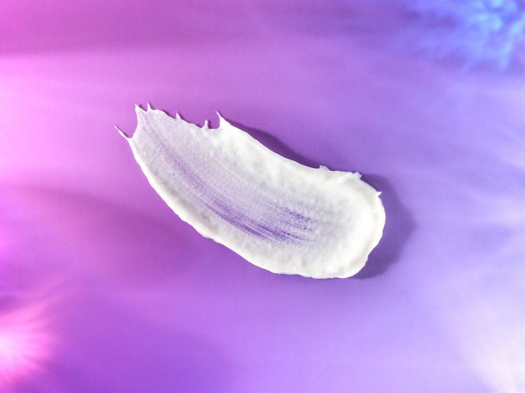 Close-up of a smear of white cream on a purple surface, accented by pink and blue light effects, showcasing texture and skincare.