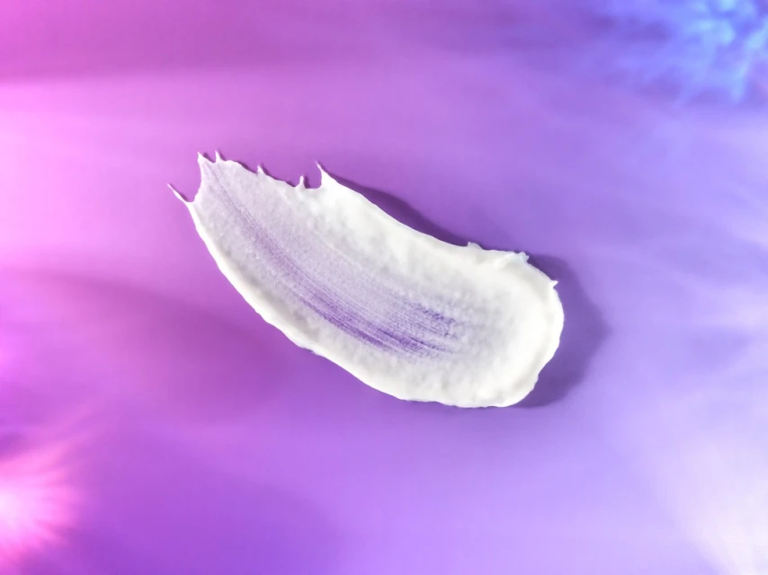 Close-up of a smear of white cream on a purple surface, accented by pink and blue light effects, showcasing texture and skincare.