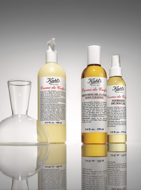 Three Kiehl's Creme de Corps body care product bottles of varying sizes are arranged with a glass decanter against a gradient gray background.