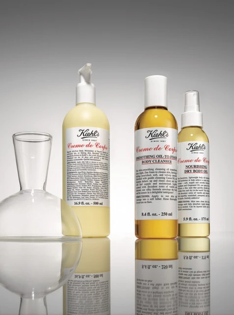 Three Kiehl's Creme de Corps body care product bottles of varying sizes are arranged with a glass decanter against a gradient gray background.