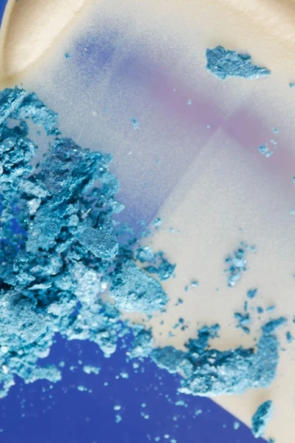 Close-up of scattered turquoise eye shadow powder on a reflective surface, vibrant blue background, beauty product.