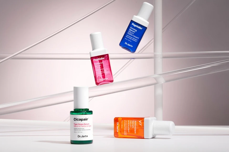 Four Dr. Jart+ serum bottles in green, red, orange and blue colors are displayed against a gradient light-pink background with clear rods.