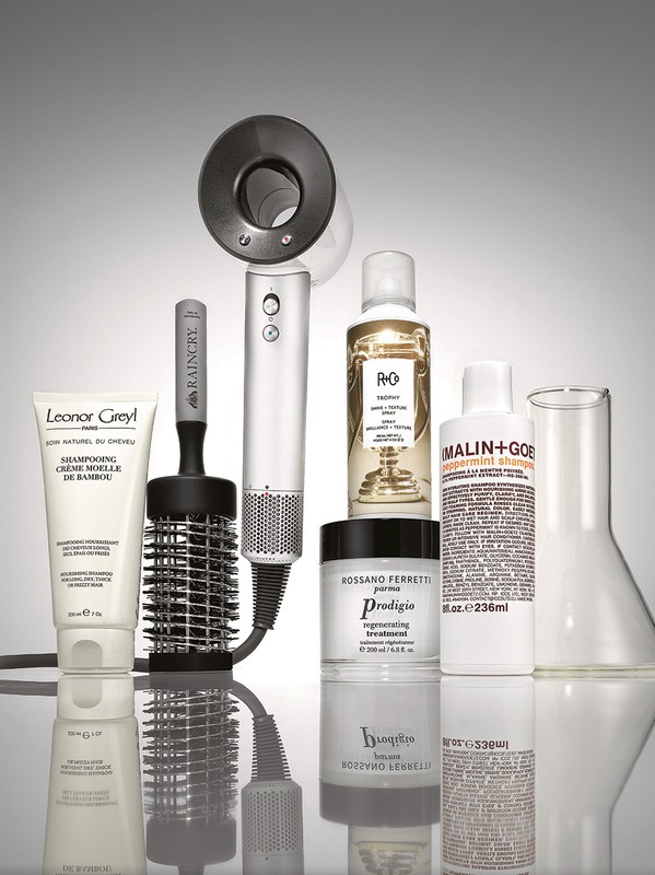 Still life of Leonor Greyl shampoo, Raincry hairbrush, R+Co, Rossano Ferretti, and Malin+Goetz hair products, and glass beaker on a reflective surface.