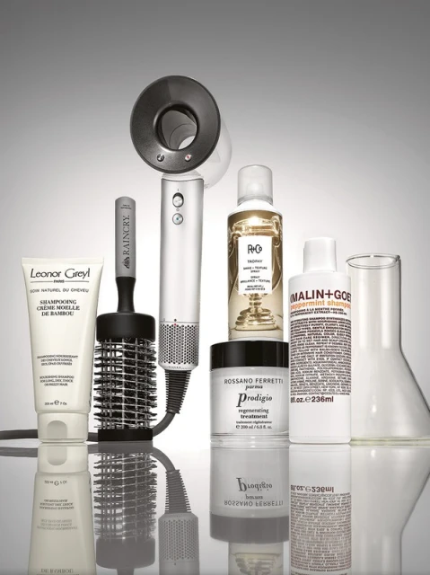 Still life of Leonor Greyl shampoo, Raincry hairbrush, R+Co, Rossano Ferretti, and Malin+Goetz hair products, and glass beaker on a reflective surface.