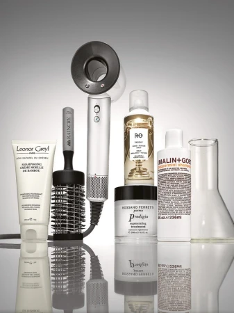 Still life of Leonor Greyl shampoo, Raincry hairbrush, R+Co, Rossano Ferretti, and Malin+Goetz hair products, and glass beaker on a reflective surface.
