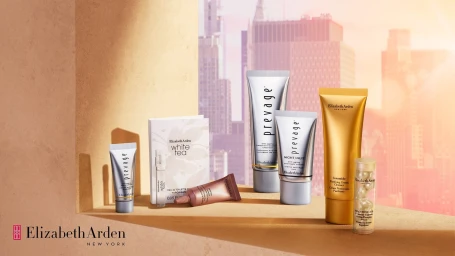 Collection of Elizabeth Arden skincare and fragrance products arranged on a tan podium against a city skyline at sunset.