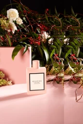 Gucci perfume bottle styled with white roses and dark foliage on a pink surface