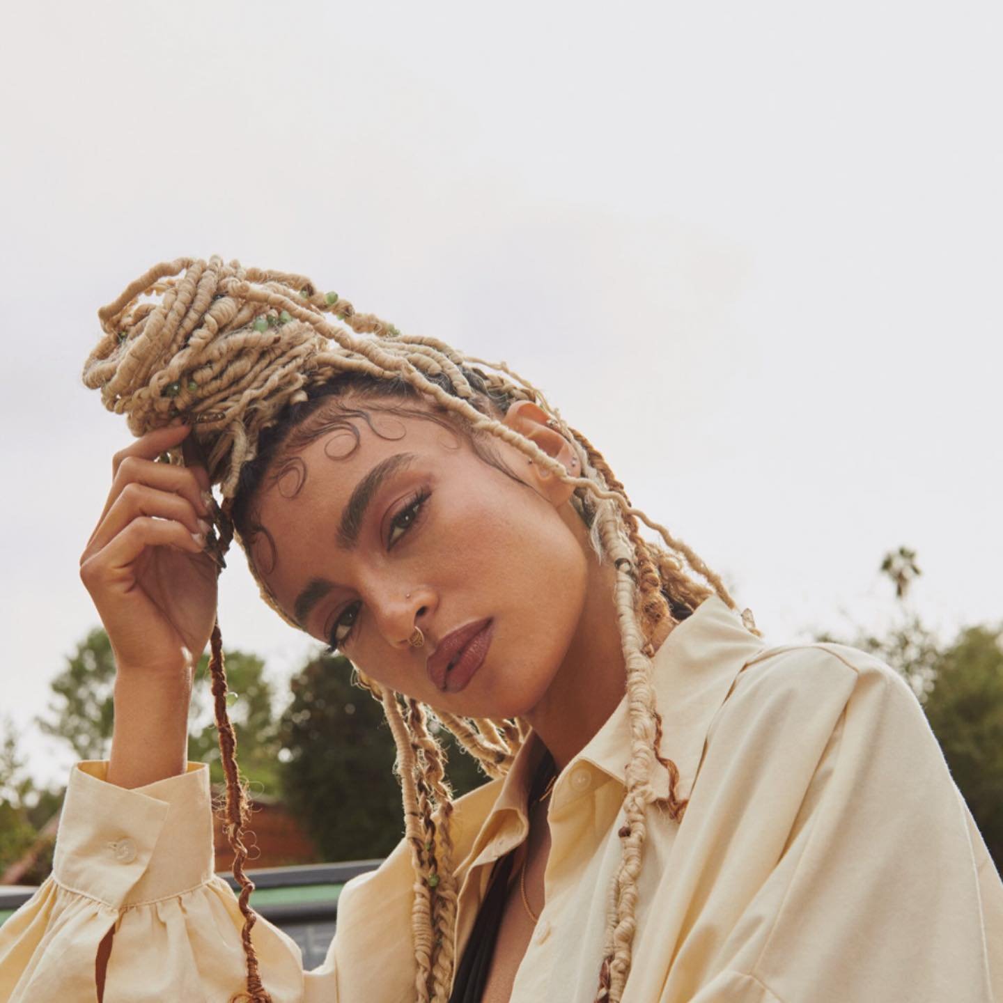 Portrait of a young woman with blonde dreadlocks, wearing a beige shirt, looking downward against a blurred outdoor background.