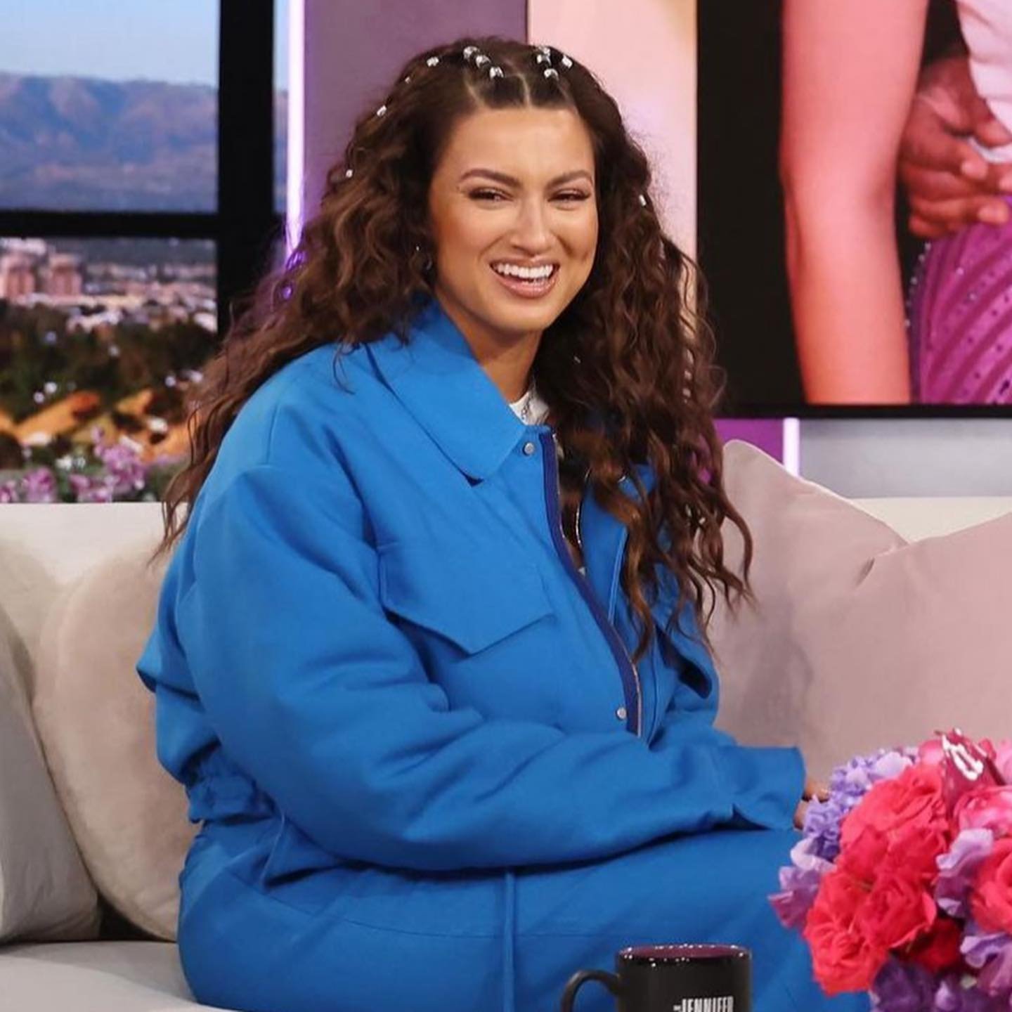 Smiling woman with curly hair wearing a blue jumpsuit is sitting on a sofa indoors, with a city view visible through a window.