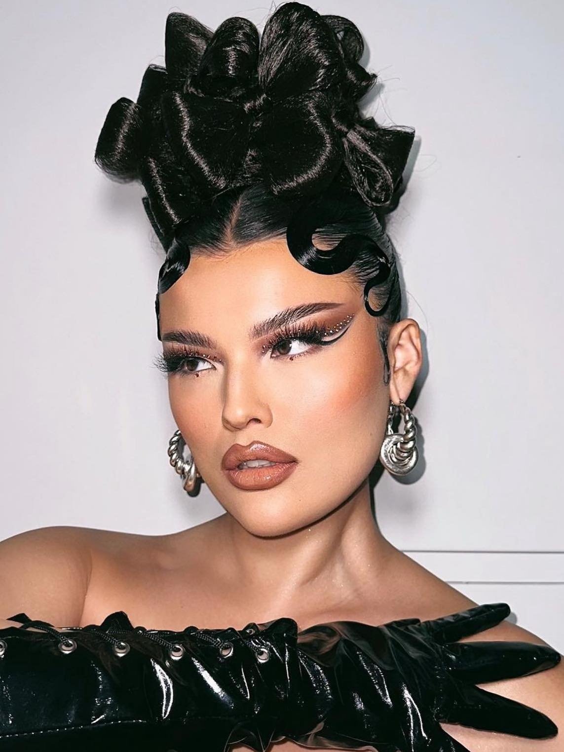 Close-up of a woman with elaborate black hair, dramatic makeup, silver earrings, and black gloves, set against a bright background.