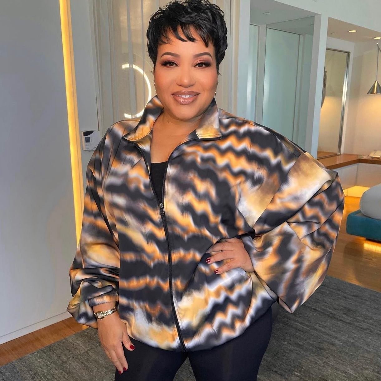 Woman wearing a colorful patterned jacket stands indoors with a neutral expression in a medium shot with soft lighting.