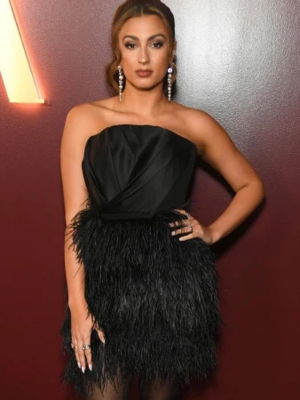 Elegant woman in strapless black dress with feathered skirt, posing indoors against a dark red wall with soft, diffused lighting.