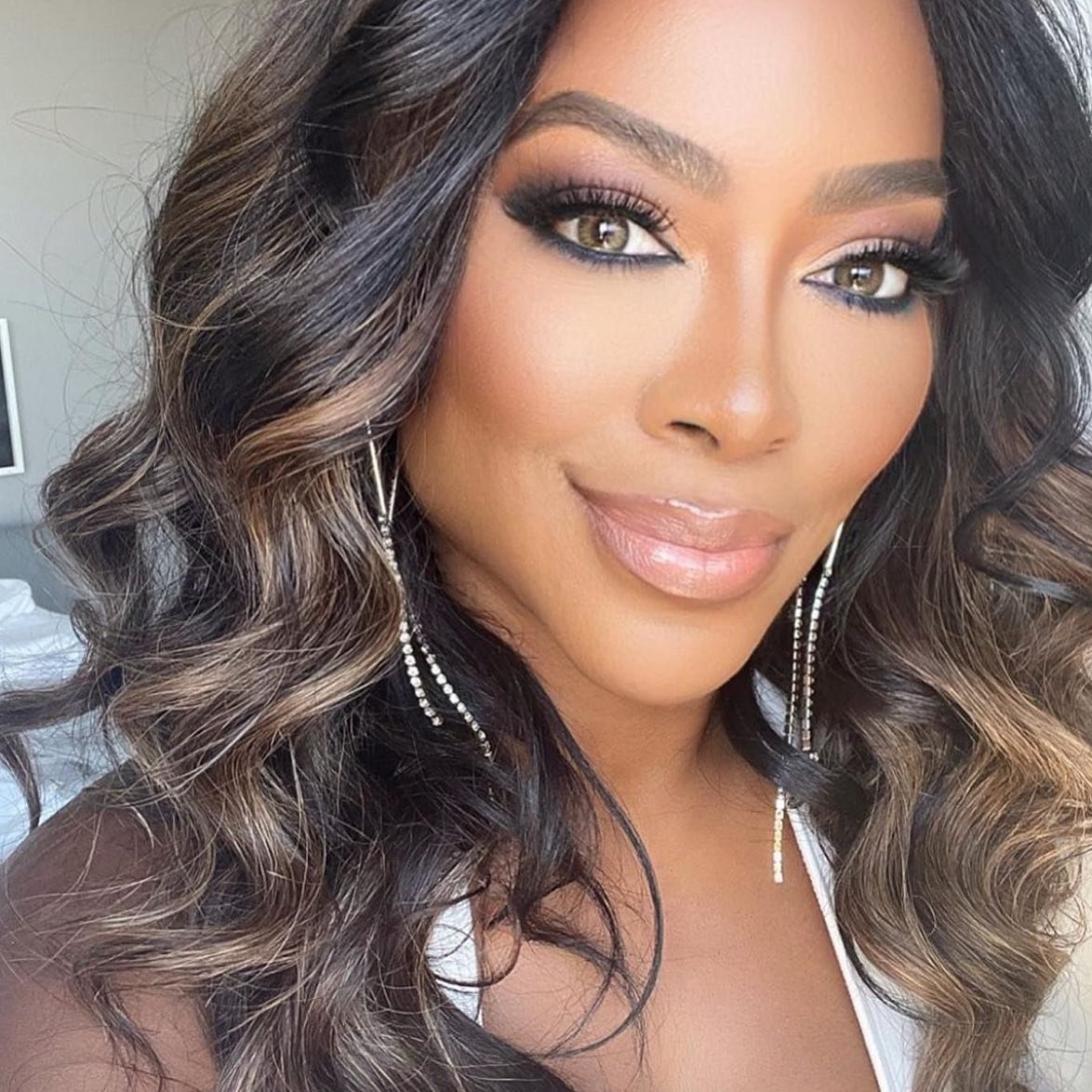 Close-up portrait of a woman with warm-toned skin, dark wavy hair, and dramatic makeup, set against a blurred neutral indoor backdrop.