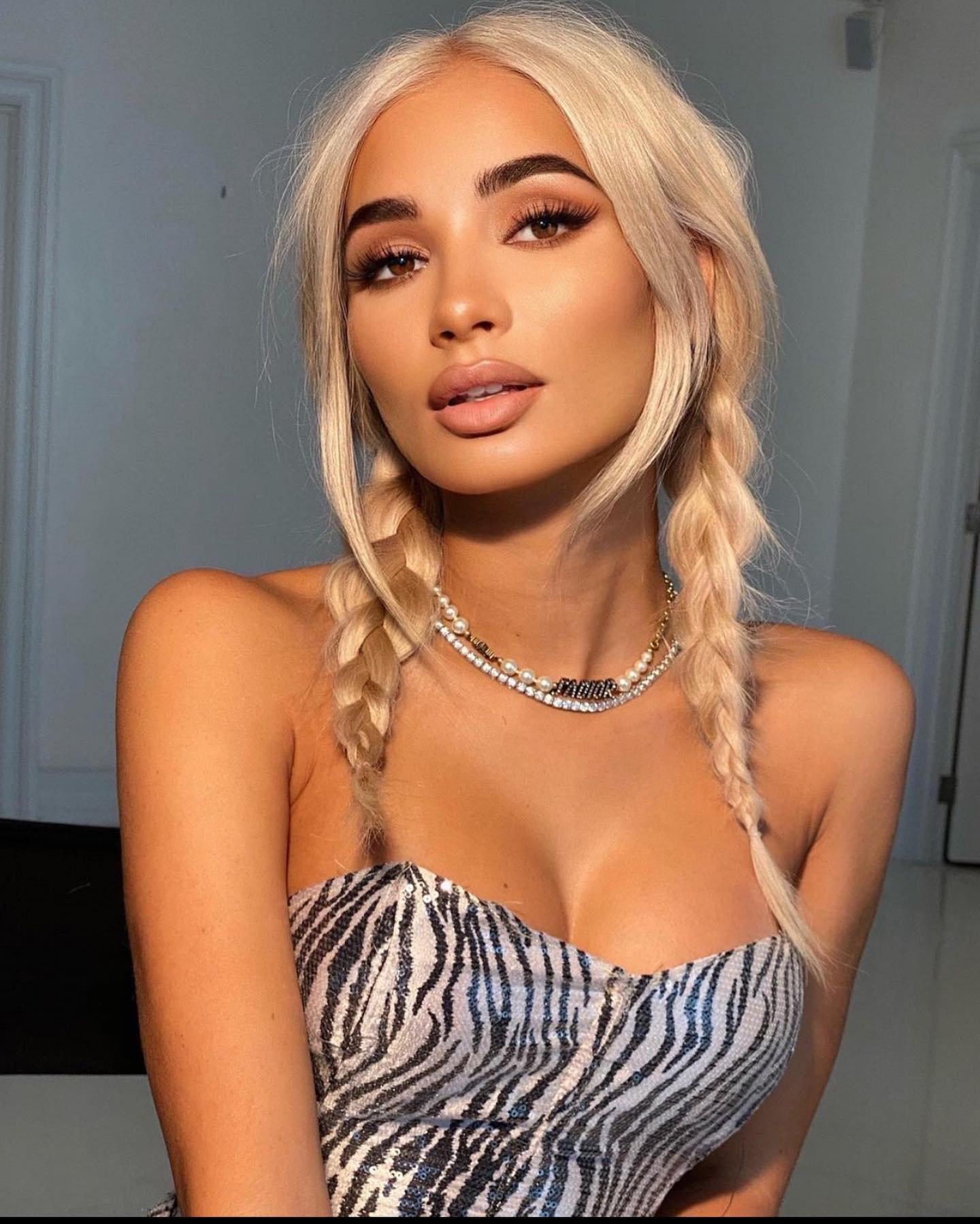 Portrait of a young woman with braided blonde hair, wearing a zebra-striped top and layered pearl and gold necklaces in a studio setting.