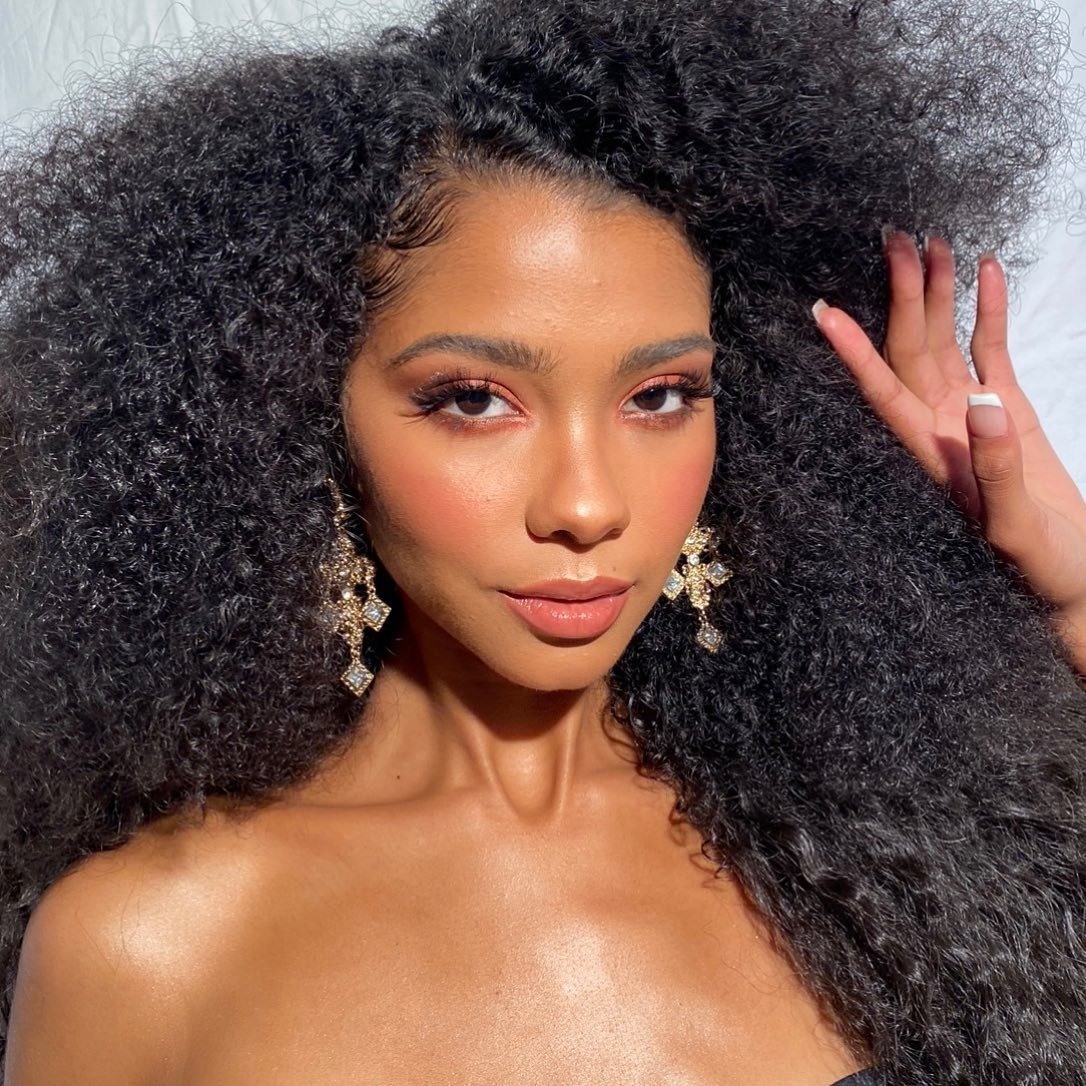 Glamorous close-up of a woman with dark curly hair, wearing chandelier earrings and makeup, with a hand touching her hair.