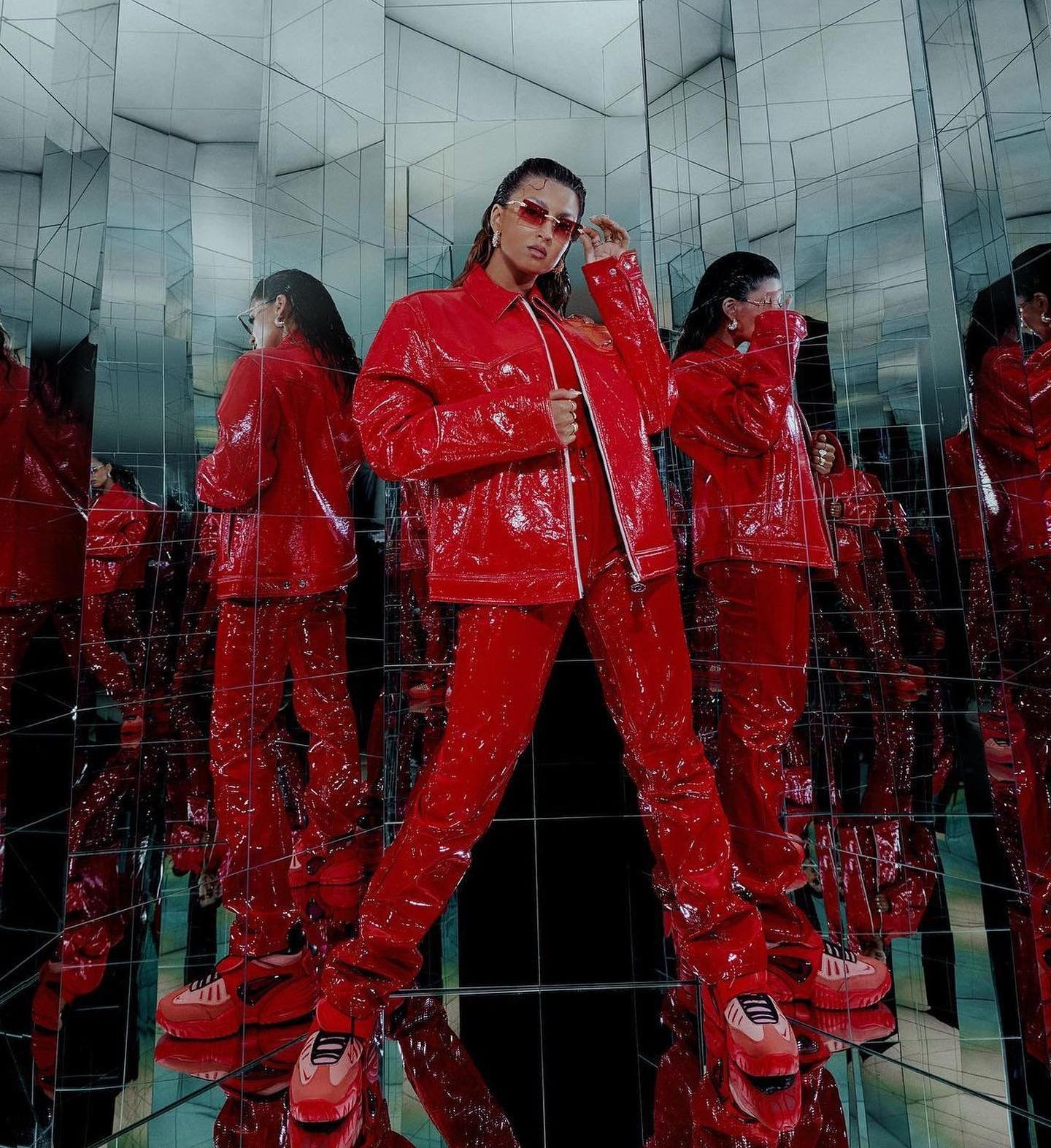 A woman in a shiny red vinyl outfit and matching sunglasses poses in a mirrored room, creating a dynamic and stylish fashion statement.