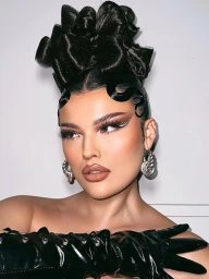 Close-up portrait of a woman with elaborate black sculpted updo hair, graphic winged eyeliner, and silver hoop earrings against a white wall.