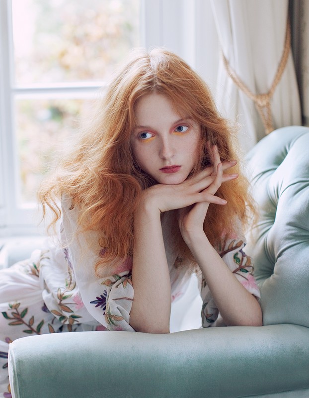 Red-haired woman rests on a blue tufted chaise lounge by a window. She wears a floral-print robe, with her hands clasped by her face.