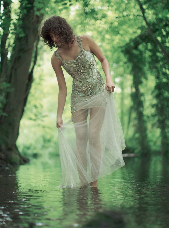 Woman in embellished dress wading in shallow water of a stream in a lush green forest. Tranquil nature scene with dreamy ethereal light.