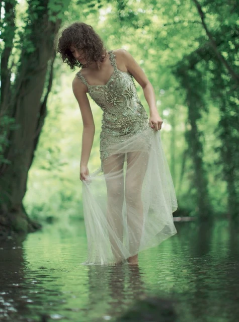 Woman in embellished dress wading in shallow water of a stream in a lush green forest. Tranquil nature scene with dreamy ethereal light.