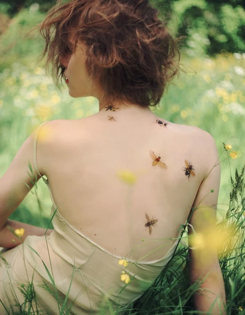 A woman sits in a field, her back to the camera, with several non-discernible bugs on her back. She has short curly hair and is wearing a light dress.
