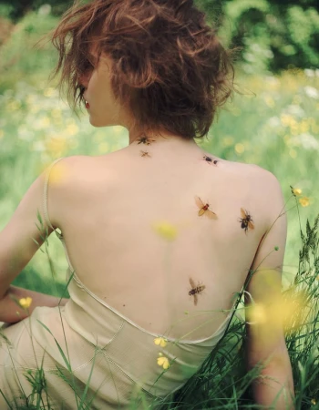 A woman sits in a field, her back to the camera, with several non-discernible bugs on her back. She has short curly hair and is wearing a light dress.