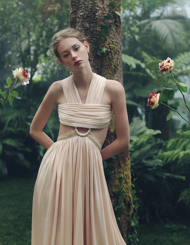 A young woman poses in a flowing dress beside a tree in a lush garden setting with roses, showcasing a fashion-forward, natural aesthetic.