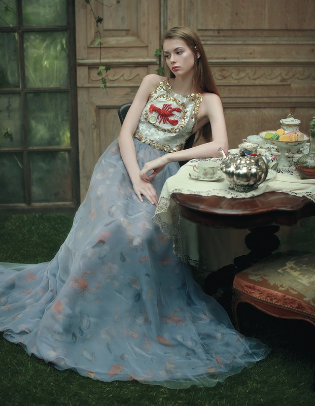 Woman in formal gown with lobster design sits at antique table with tea set in garden setting with vintage architectural details.