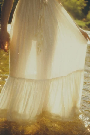 Close-up of a person wearing a sheer white maxi dress standing in shallow natural water under warm sunlight.