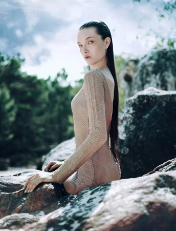 Woman with long dark wet-look hair wearing a nude textured dress seated on rock formations outdoors under a cloudy sky.