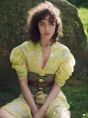 Woman with curly dark hair wearing a yellow floral dress and wide laced corset seated outdoors near a mossy rock.