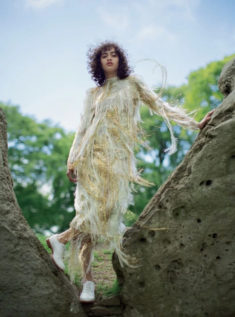 Woman in fringed dress posing on rocks outdoors, with curly hair, white shoes, against a blurred natural background, fashion editorial.