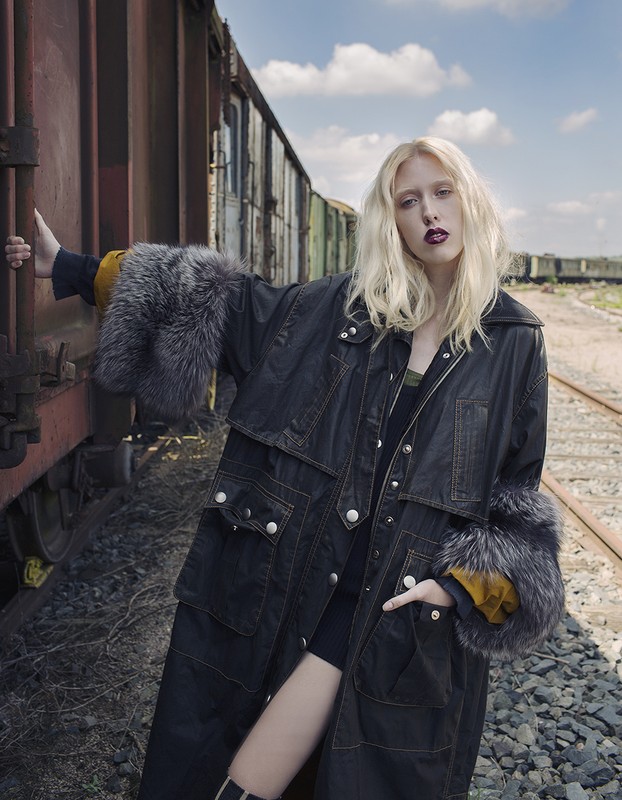 A woman with fair hair and a black jacket poses against a backdrop of train cars and tracks in an outdoor environment.