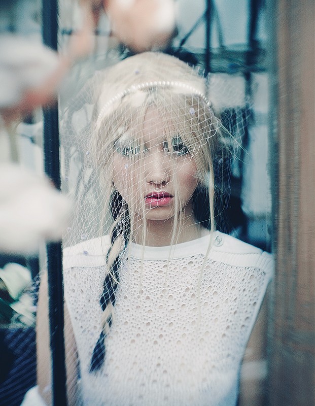 Portrait of a young woman with blond hair and pink lipstick wearing a white, sleeveless top and a beaded net head covering.