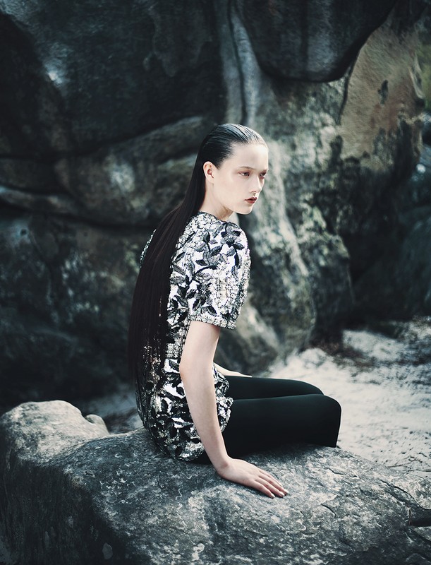 A stylish woman with slicked-back hair poses while seated on a rock amidst a rocky landscape. She wears a shiny patterned top and leggings.