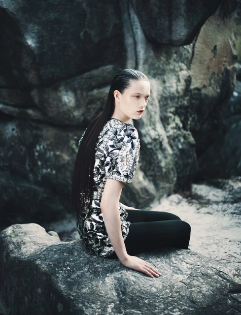 A stylish woman with slicked-back hair poses while seated on a rock amidst a rocky landscape. She wears a shiny patterned top and leggings.