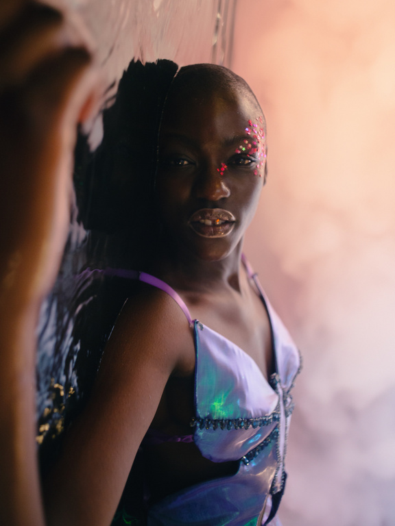Young woman with glitter makeup poses indoors, wearing a shiny iridescent top with black trim, reflecting light in a soft studio.