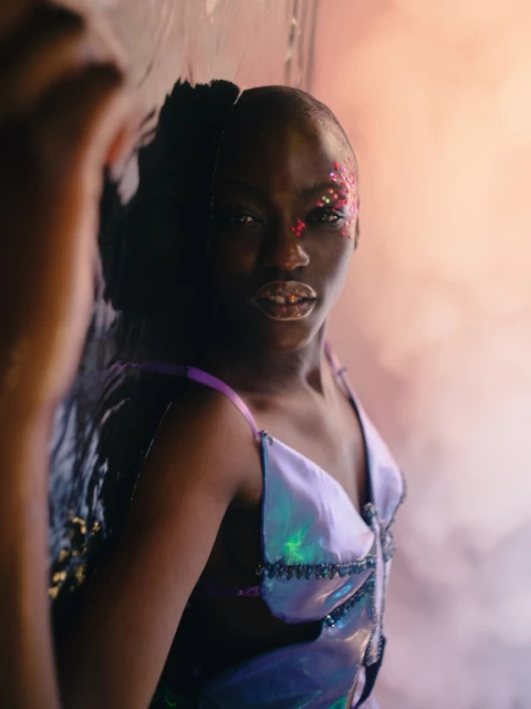 Young woman with glitter makeup poses indoors, wearing a shiny iridescent top with black trim, reflecting light in a soft studio.