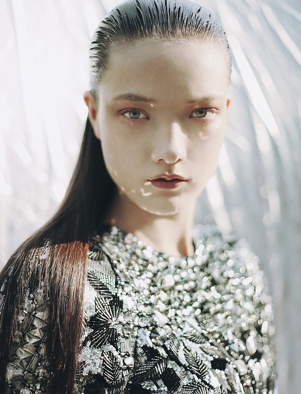 Close-up portrait of a young adult woman with slicked-back hair and a silver sequined top against a blurred silver background.