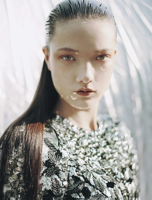 Close-up portrait of a young adult woman with slicked-back hair and a silver sequined top against a blurred silver background.