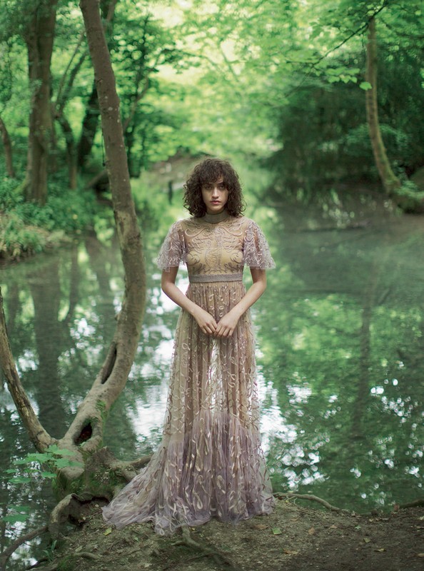 Young woman in a formal gown stands gracefully by a tranquil river surrounded by lush trees in a vibrant, nature-filled environment.