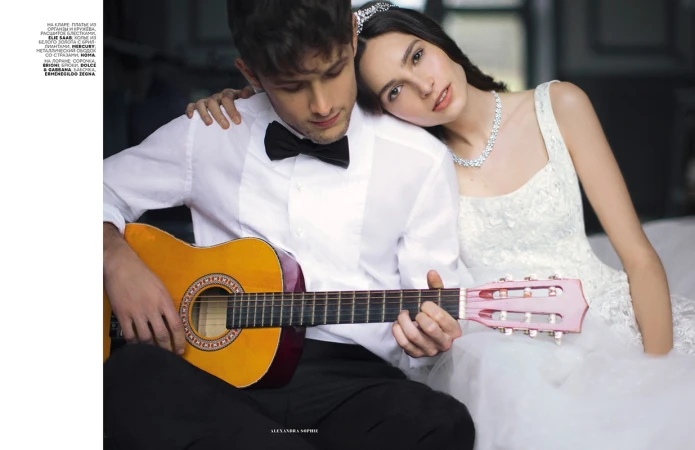 Elegant couple in formal wear. Woman in bridal gown rests head on man's shoulder as he plays guitar in a soft, romantic indoor setting.