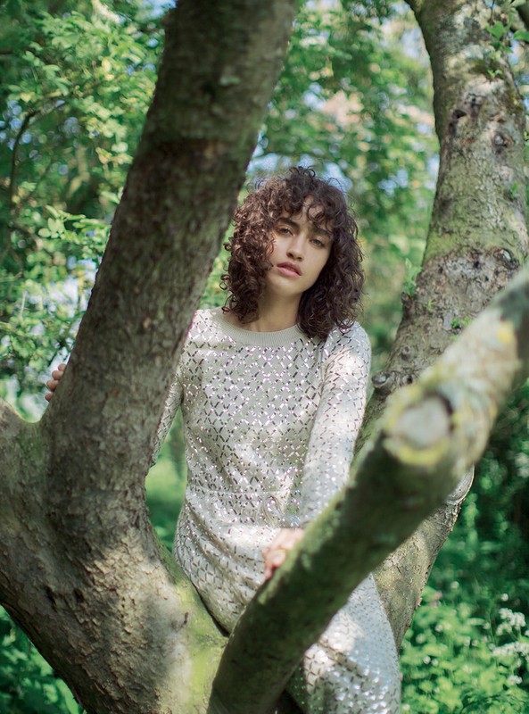 Adult female model with curly hair poses in a sparkly dress, sitting in a tree surrounded by green leaves on a sunny day.