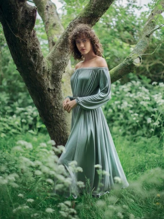 Woman in an off-the-shoulder grey dress standing among trees and wildflowers in a lush woodland setting photographed by Alexandra Sophie