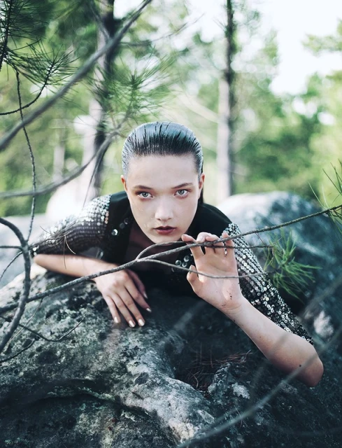 A stylish woman with slicked-back hair is the primary subject, captured outdoors with a blurred green forest backdrop. She poses on a gray stone.