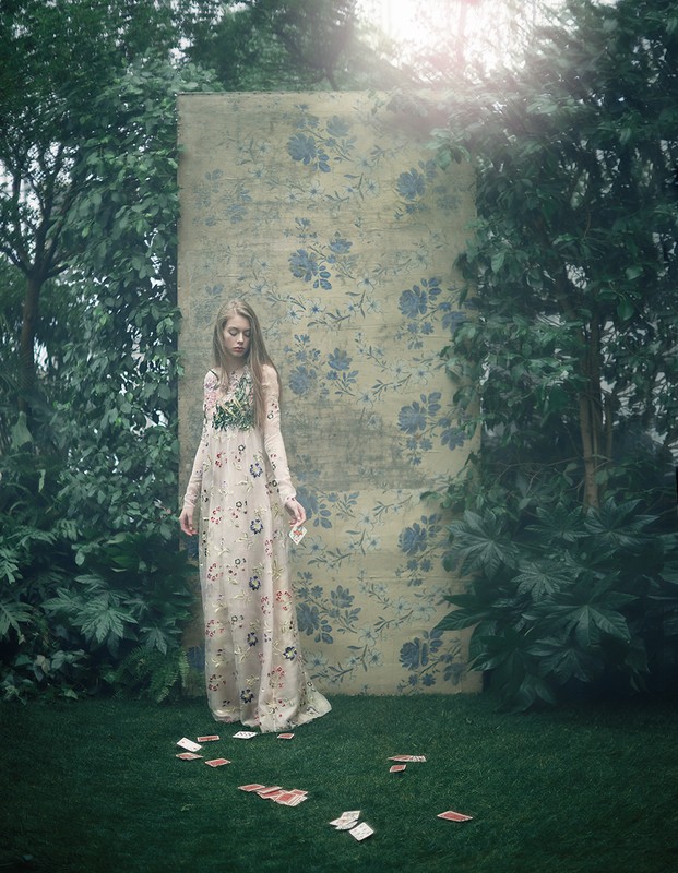 A woman in a floral dress stands among playing cards on a lawn, framed by lush greenery and a floral backdrop, creating a serene outdoor scene.