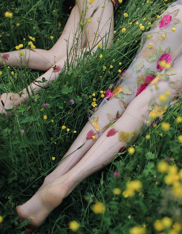Close-up of two women in floral dresses lying in a field of yellow wildflowers and green grass in natural, soft lighting.