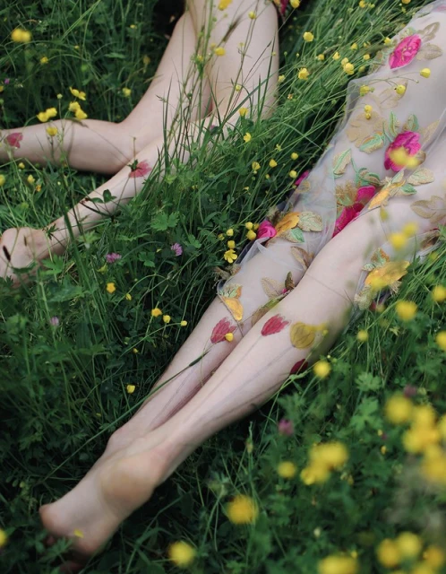 Close-up of two women in floral dresses lying in a field of yellow wildflowers and green grass in natural, soft lighting.