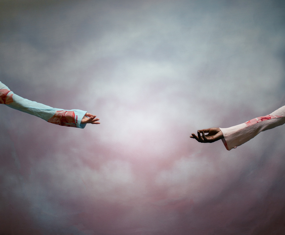 Two arms reach toward each other with open hands against a backdrop of diffused pink, white, and grey clouds, creating an ethereal scene.